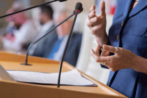 Close-up of unrecognizable female politician in rings standing at rostrum with microphones and clipboard while addressing conference