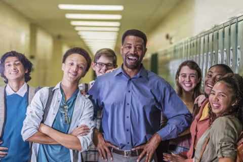 A multi-ethnic group of six high school students hanging out together in the hallway with a teacher smiling